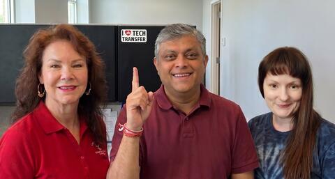 From left to right, Cecelia Foote, Saikat Kumar Ghosh, and J'aime Maynard smiling and posing for a group photo in an office, with Saikat Kumar Ghosh pointing to a "I (Heart) Tech Transfer" Sign in the background behind them. 