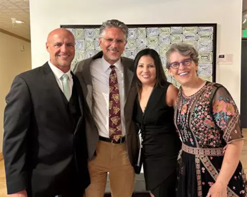 From left to right, Bill Clark, Dan Dender, Julie Leonard, and Jody Sandel pose together in formal attire, with a framed piece of artwork and a small descriptive display card behind them.