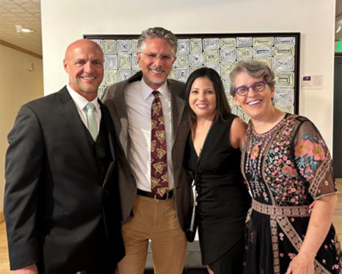 From left to right, Bill Clark, Dan Dender, Julie Leonard, and Jody Sandel pose together in formal attire, with a framed piece of artwork and a small descriptive display card behind them.