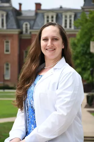 Portrait picture of Korina Menking-Hoggatt,Ph.D, in front of the brick building Wilburn Hall in a lab coat 