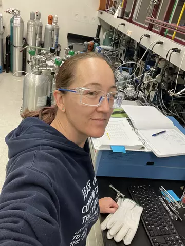 Woman wearing safety glasses standing at lab bench with gas cylinders in background.