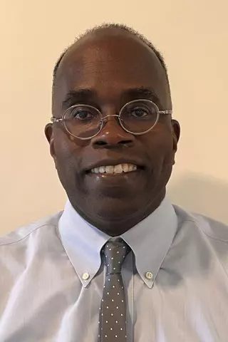 Bryant C. Nelson wearing glasses, a light gray dress shirt, and a gray patterned tie, smiling for the camera.