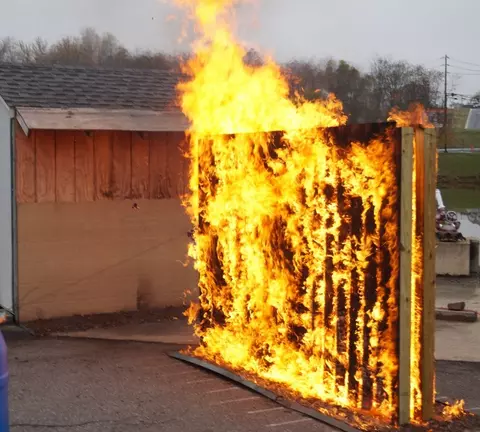 A photo of two back-to-back wood privacy fence panels spaced eight inches apart burning in front of a shed. The flames engulf the entire set of fences and extend more than three feet above them.