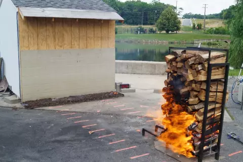 A photo of a tall and narrow stack of firewood burning in front of a shed under a wind speed of 6 meters per second or 13 miles per hour. The logs are 41 centimeters or 16 inches long and are stacked 1.22 meters or four feet high by 61 centimeters or two feet long, at a distance of 3.66 meters or twelve feet from the shed. The logs are oriented parallel to the shed. Flames extend out from the sides of the lower half of the stack.