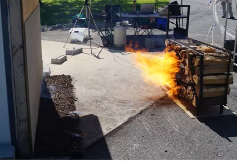 A photo of a low and wide stack of wood burning in front of a shed under a wind speed of 10 meters per second or 22 miles per hour. The logs are 16 inches long and are stacked 2 feet high by 4 feet long, at a distance of 6 feet from the shed. The logs are arranged perpendicular to the shed, so that the ends face the shed. Flames extend directly toward the shed.