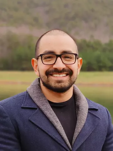 headshot of Abraham Kuri Cruz, a man with short brown hair, beard, and glasses, wearing a blue blazer, smiling outdoors.