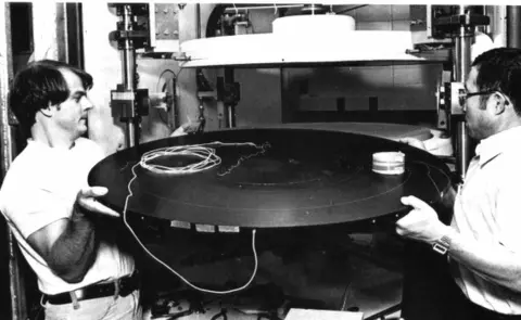 Two men with a large circular object. black & white
