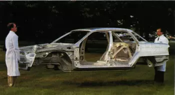 Two researchers in lab coats hold a sedan's aluminum frame off the ground in a grass field.