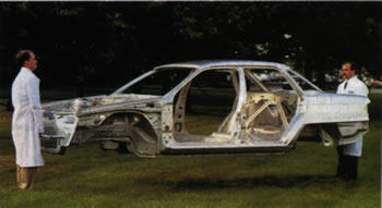 Image of two men in lab coats holding a sedan's aluminum frame off the ground in a grass field.