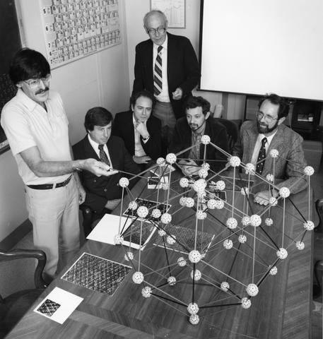 a group of men in a room with a large model of a crystal. One of the men (Shechtman) points at a section of the crystal.