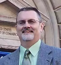 Headshot of Vincent A. Hackley, a man with short hair, glasses, and beard, smiling, wearing a suit and tie in front of a government building.