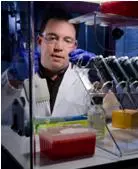 Kevin Kiesler wears a lab coat and goggles while working at a lab bench. 