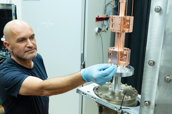 A man wearing latex gloves reaches out to adjust something on a cylindrical metal scientific device.
