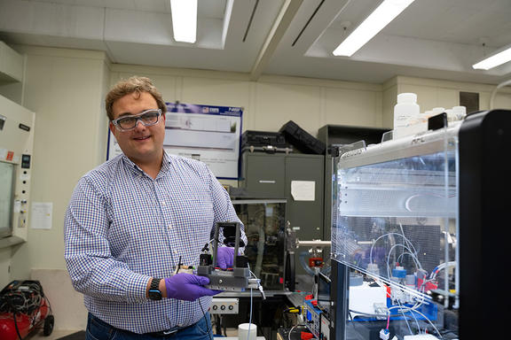 Peter Beaucage wears safety glasses as he stands in the lab, holding a piece of equipment on display. 