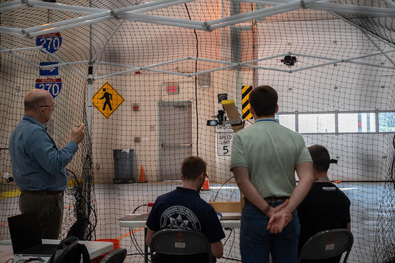 Four men are protected by a net structure as they look away from the camera toward a drone flying in a large high-ceilinged room with street signs on the walls.