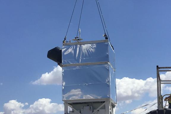 A rectangular box coated with reflective foil hangs from cables over a parking lot.