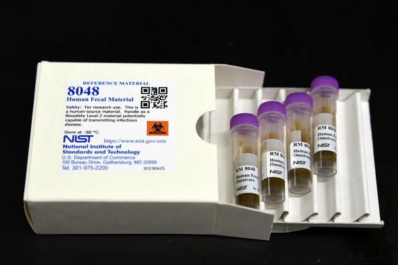 A white box labeled "8048 Human Fecal Material" lies open, with a tray of four plastic vials pulled out. 