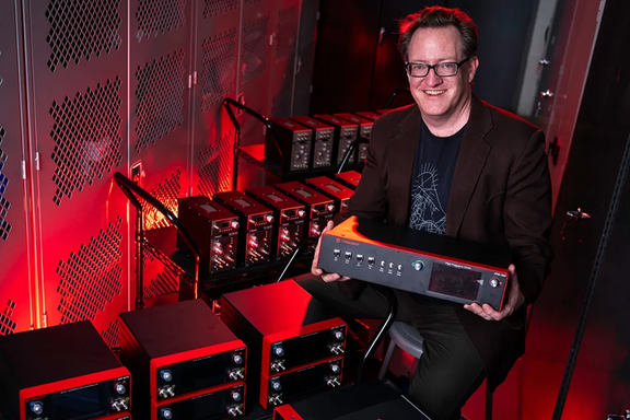 Scott Davis poses smiling and holding a frequency comb console. Other similar electronic products are stacked on carts around him. 