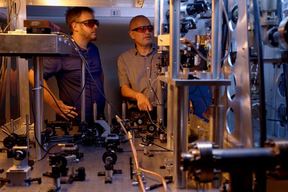 Two researchers in safety glasses stand behind a table holding various electronic and laser devices. A metal structure in the foreground is part of the atomic clock. 