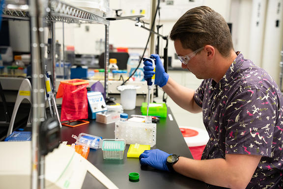 Researcher in safety glasses and gloves sits at a lab bench working with small test tubes.