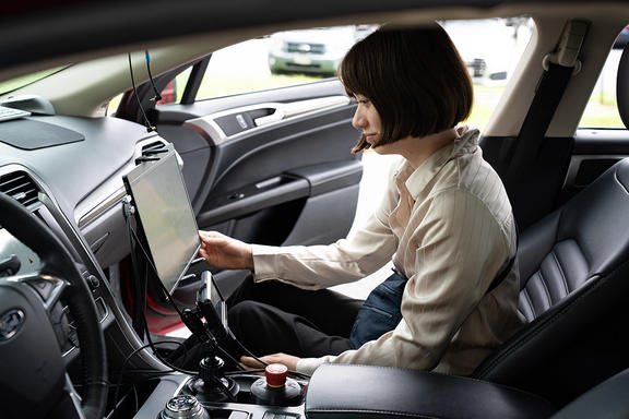 A woman sits in the passenger side of a parked car, looking at a large monitor attached to the dashboard. 