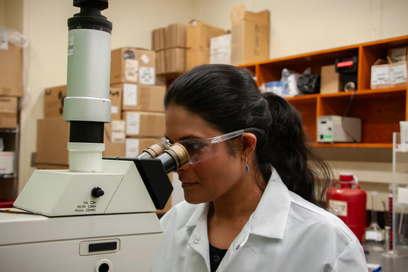 A woman wearing a lab coat and safety glasses stares into a large microscope.