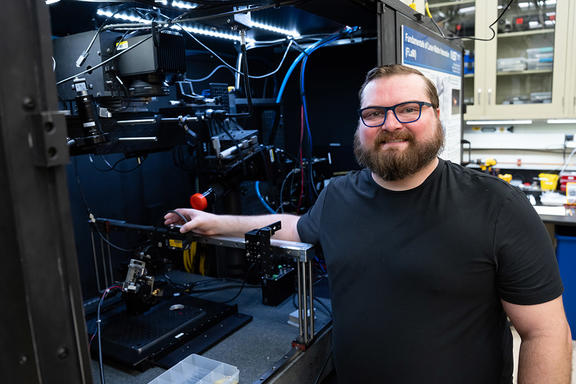 A bearded man poses smiling next to an open cabinet of equipment in a lab. 