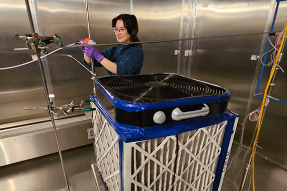 A large cube made from a box fan, pleated air filters, and duct tape sits on a table in the middle of a large air tight steel room. In the background, a scientist clamps a narrow, flexible hose in place.