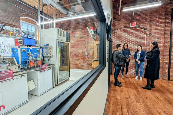 Four people stand talking in a converted factory space near a large interior window through which a complex, high-tech manufacturing line is visible. 