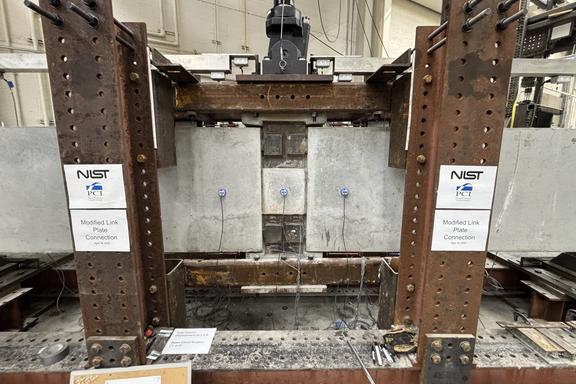A wide view of a concrete beam assembly inside a large metal frame. A hydraulic arm is part of the metal frame, and it’s posed to press down at a section in the middle where the concrete pieces connect.