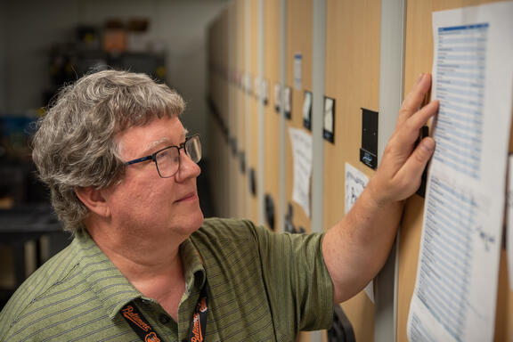Doug White stands looking at a printed-out list taped to the end of a library shelf. 