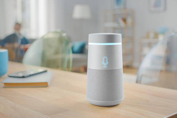 A cylindrical smart speaker sits on a wooden table in a living room. 