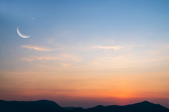 A sliver of moon is visible in the sky during sunset over mountains.