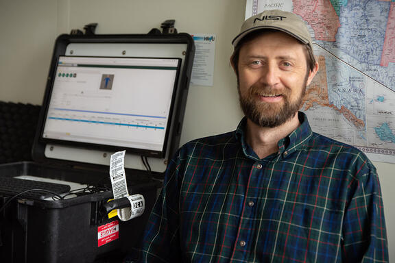Gary Howarth poses for a photo in his office, wearing a NIST baseball cap, with a ruggedized laptop on the counter next to him and a map on the wall behind him.. 