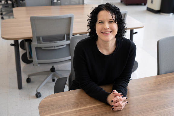 Kelly Sauerwein poses sitting at a table in the NIST library, leaning forward and smiling. 