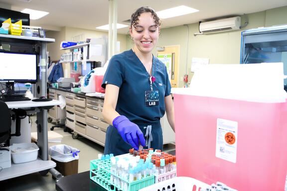 A medical worker in scrubs places a vial into a rack in a medical lab. 