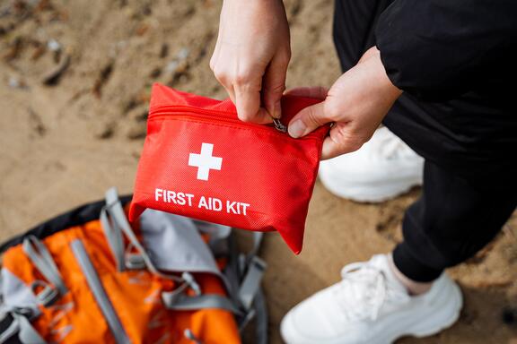 A person holds a red pouch labeled First Aid Kit as they stand over a travel bag on sandy ground.