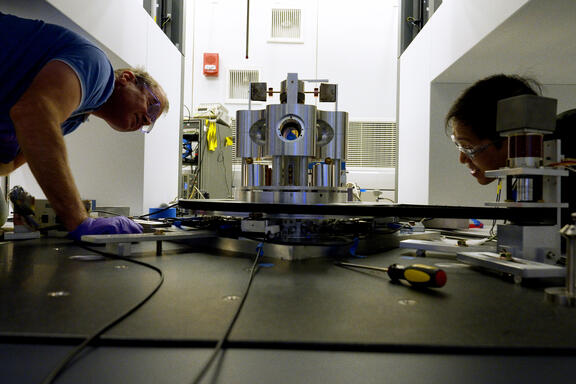 Wide shot shows two researchers on either side of a scientific device made of metal cylinders, peering closely at it. 