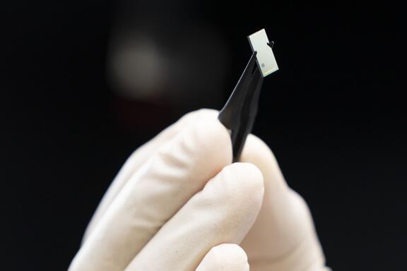 Close-up of a gloved hand holding tweezers that are grasping a tiny rectangular chip. 