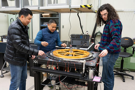 Three warmly dressed researchers stand around a scientific device with a large circular piece lying flat on top. 