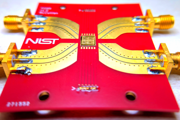A bright-red rectangle with a NIST logo holds a gold-colored chip in the center with metal paths leading to connectors at the edge of the rectangle. 