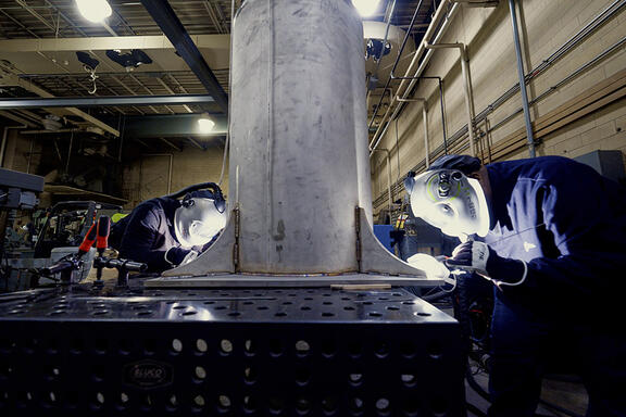 Two people wearing welding masks and other safety equipment bend over a table to work on a large metal cylinder.