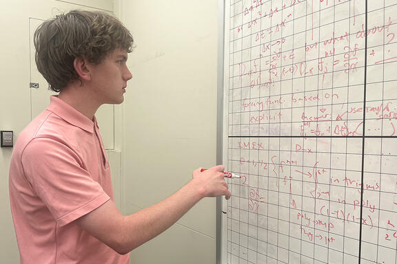 Henry Nonnemaker stands holding a marker, looking at a gridded whiteboard covered with mathematical formulas.