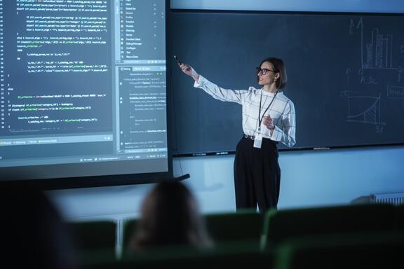 A female instructor wearing glasses presents code on a large screen to a classroom.