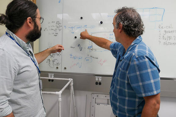 Two men face away from the camera, pointing to diagrams written on a whiteboard. 