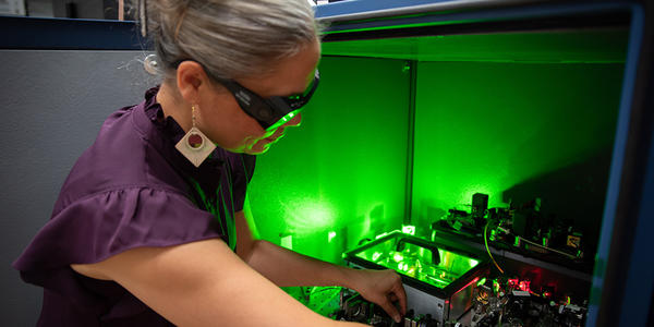 A researcher wearing safety glasses reaches into a box of circuitry and other equipment, which emits a green glow. 