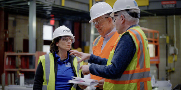 Three team members in construction safety gear stand talking inside the large warehouse where materials testing is taking place.