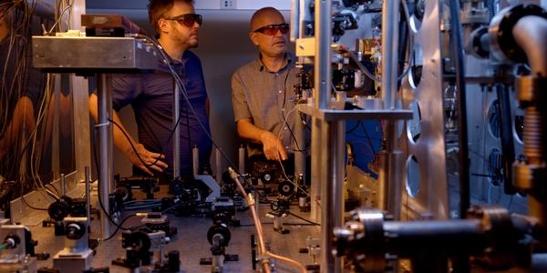 Two researchers in safety glasses stand behind a table holding various electronic and laser devices. A metal structure in the foreground is part of the atomic clock. 