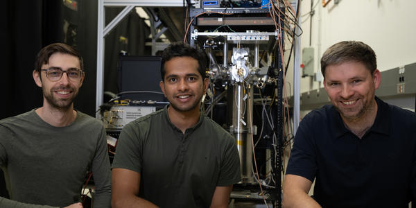 Three male researchers pose smiling in front of a large, complex scientific device in a lab.