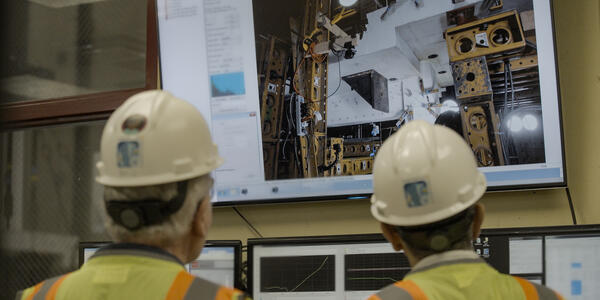 Two people in hard hats, shown from behind, look at a large screen showing concrete slabs and testing equipment.